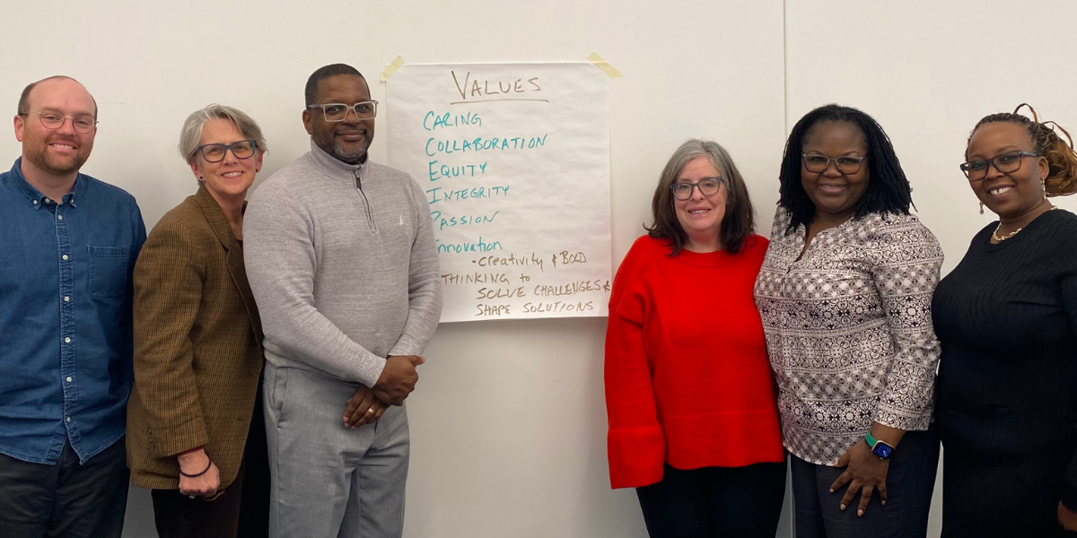 Six members of AfriThrive's Infrastructure Taskforce stand together at their inaugural meeting on January 29, 2026, next to a flip chart listing the group's shared values: Caring, Collaboration, Equity, Integrity, Passion, Innovation, Creativity and Bold Thinking to Solve Challenges and Shape Solutions